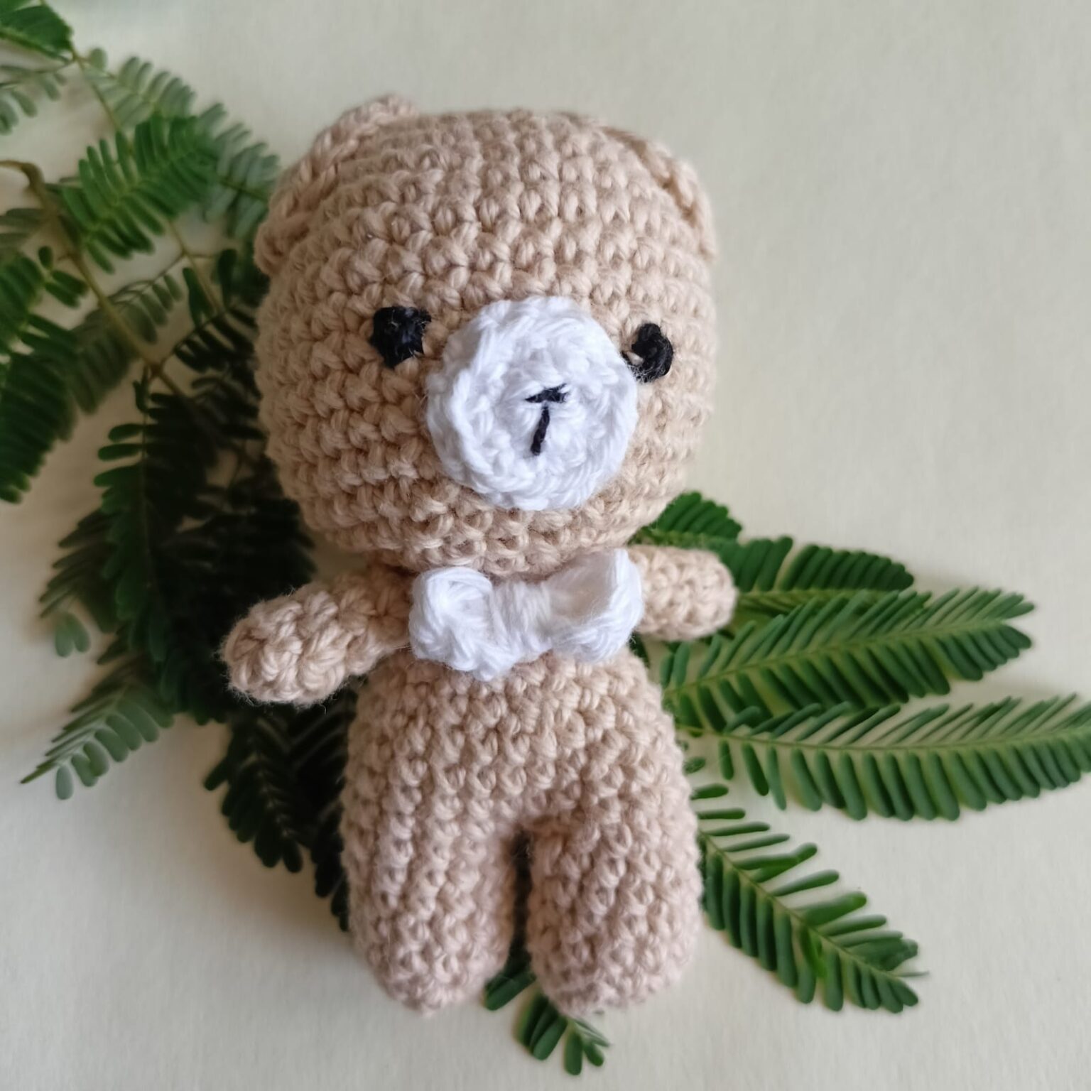 Handmade crochet teddy bear keychain with a bow on a green leafy background